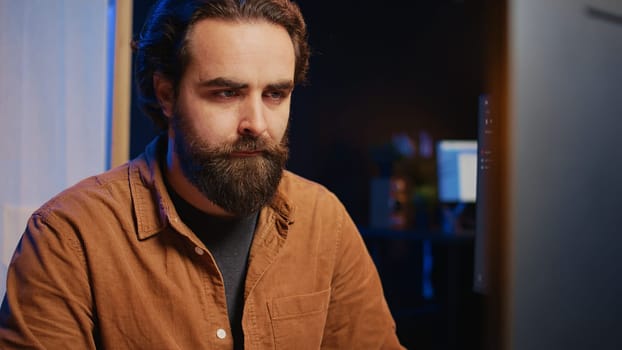 Software technician in apartment office deeply immersed in coding on computer at desk, close up. IT freelancer absorbed by work, focused on screen, writing lines of code at workstation, camera B