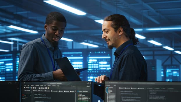 Coworking system administrators in server hub monitoring data center energy consumption across hardware using tablet. Colleagues inspecting supercomputers, ensuring malfunctions are fixed