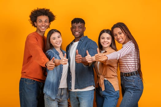 A multiracial group of five young adults stand in front of a yellow backdrop, each giving a thumbs up gesture. They are all smiling and appear to be in good spirits.