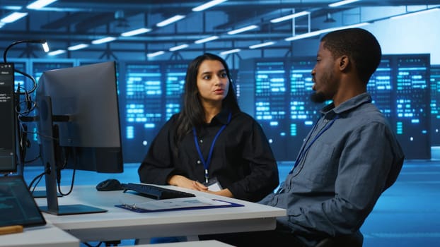 Multiracial team of IT engineers in server room brainstorming ways to fix equipment storing datasets. Indian and african american coworkers discussing how to mend racks doing computational operations