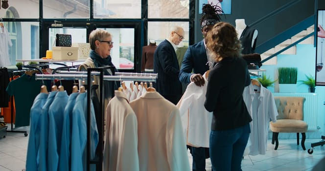 Fashion stylist suggesting clothing items to regular customer, team of store workers help elderly woman to buy trendy clothes on sale. Retail clerks assisting client during shopping spree. Camera B.