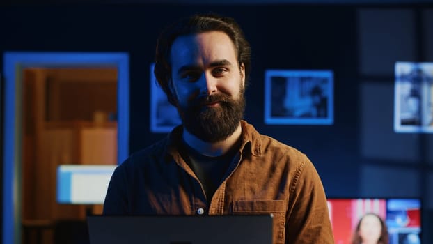 Portrait of happy man doing IT support job from apartment, standing in personal office with laptop in hand, coding. Joyful developer writing lines of code on notebook, camera A close up shot