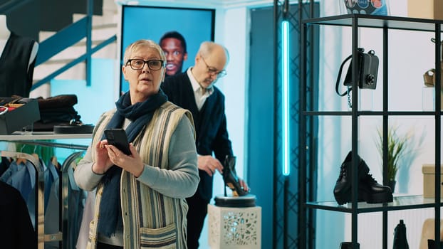 Elderly woman checking website stock to find items in store at mall, shopping for the new designer collection. Customer using smartphone app and looking for formal clothing line. Camera A.