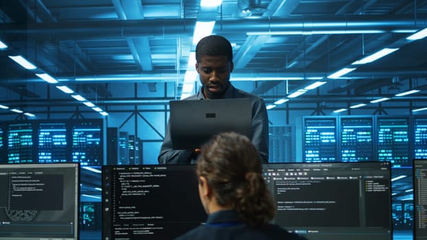 Smiling african american manager in data center doing checkup on electronics to avoid downtime. Team leader holding laptop supervising employees in server room infrastructure components