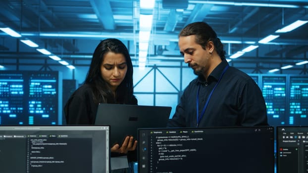 Technicians working in high tech server hub, analyzing data on laptop to ensure seamless data flow. Coworkers examining equipment in data center, ensuring system integrity and security