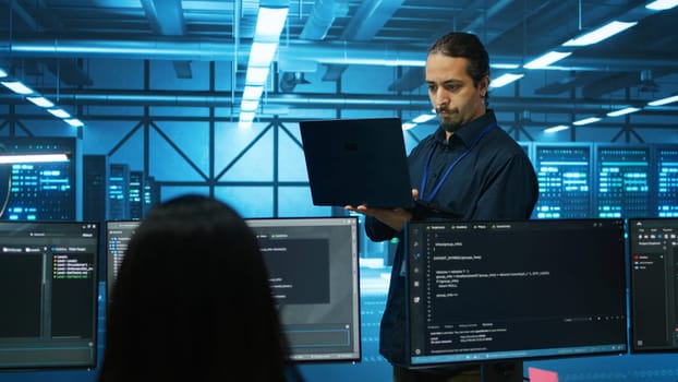 Engineer typing on laptop in server hub, checking recovery plan, monitoring data center energy consumption across components. Technician inspecting rackmounts, doing routine disk checks