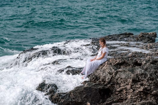 A woman in a storm sits on a stone in the sea. Dressed in a white long dress, waves crash against the rocks and white spray rises