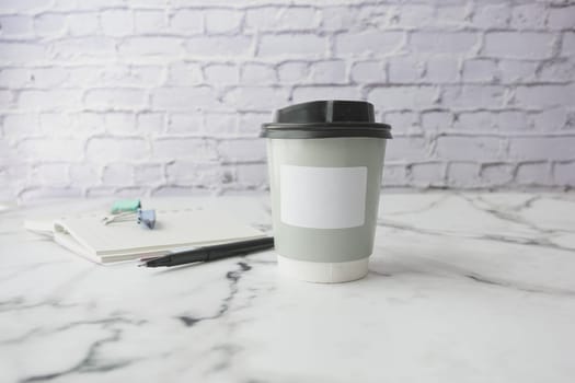 Disposable coffee cup on a marble table next to a notebook and pen, set against a brick wall background.
