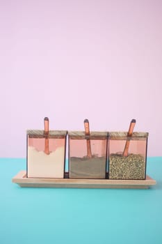 Three glass jars with different spices and copper spoons on a pastel background, wooden tray included.