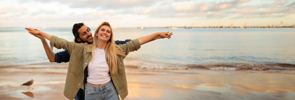 A couple enjoys the warmth of the evening sun as they stand together on a sandy beach, the woman with her arms outstretched and the man holding her from behind, panorama with copy space
