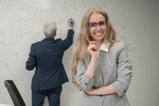 An elderly man writes on a white board and a young woman stands thoughtfully