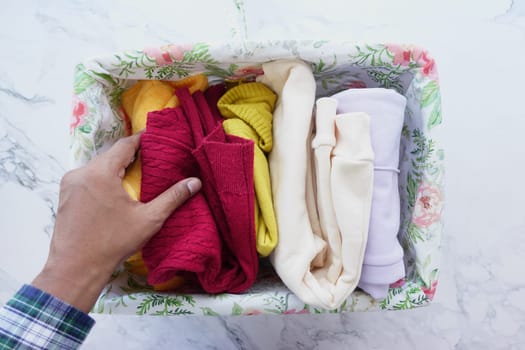 A hand arranging colorful clothes in a floral storage box on a marble surface, emphasizing organization and decor