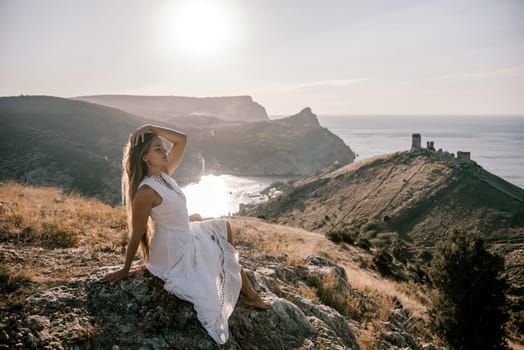 A woman in a white dress is sitting on a rock overlooking a body of water. She is enjoying the view and taking in the scenery