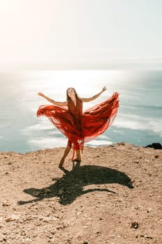 Woman red dress sea. Female dancer in a long red dress posing on a beach with rocks on sunny day. Girl on the nature on blue sky background