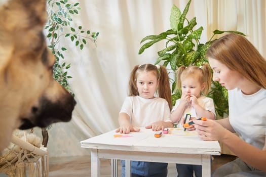 Young mother or babysitter, little daughter, sister teenager girl drawing at table and big dog shepherd. Family enjoying leisure at home