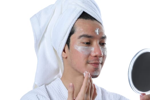Young man applying moisturizer on his face and looking at mirror over white background.