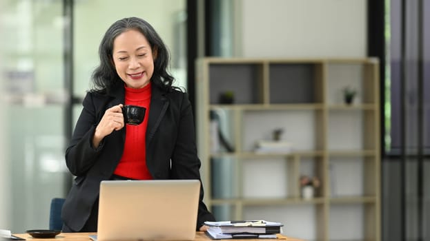 Successful mature businesswoman enjoying hot drink and using laptop at her workplace.