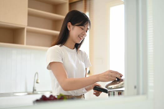 Attractive young asian woman standing by the stove, preparing lunch in modern kitchen.