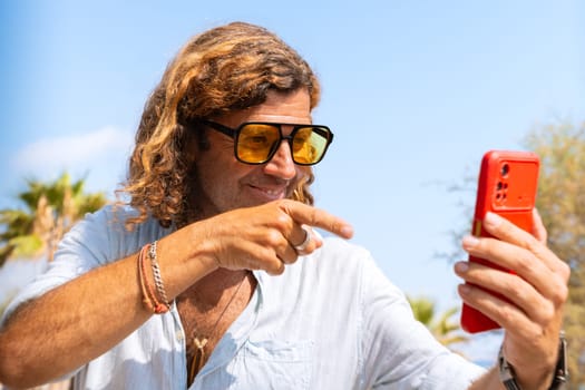 Happy mid adult man wearing sunglasses on vacation on the beach greeting his family by video call with his smartphone.