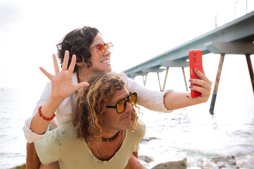 Happy mid adult couple wearing sunglasses on vacation on the beach greeting his family by video call with his smartphone.