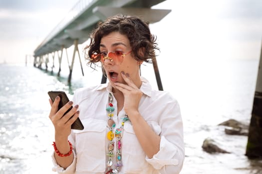 Caucasian woman wearing sunglasses vacationing by the sea with a gesture of surprise using a social media app with her mobile phone