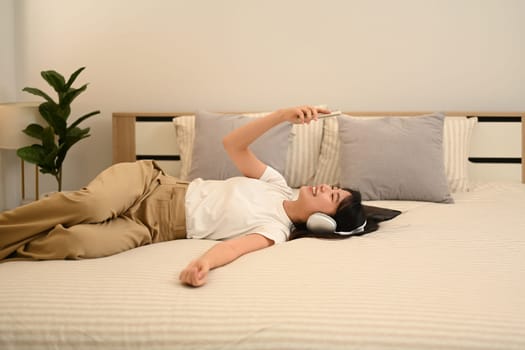 Cheerful asian woman with wireless headphones using smartphone while lying in bed.