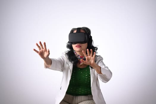 Excited middle age woman wearing VR headset and trying to touch something on white background.