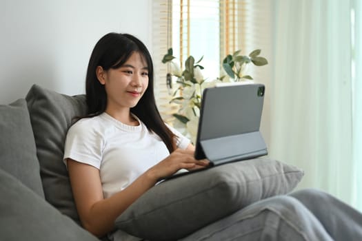 Beautiful young Asian woman relaxing on couch at home and using digital tablet.