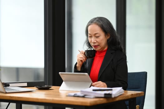 Professional senior businesswoman drinking coffee and using digital tablet at office desk.