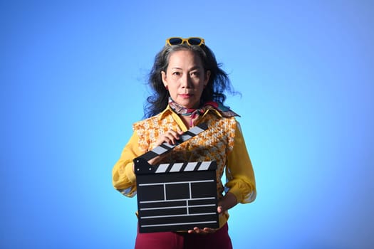 Fashionable middle age woman holding clapperboard isolated on blue background.