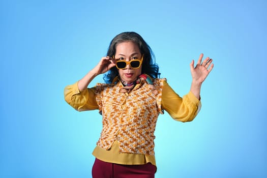 Attractive mature woman with fashionable clothes and sunglasses posing over blue background.