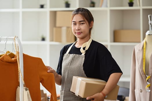 Portrait of young female small business owner holding cardboard box standing at her clothing shop.