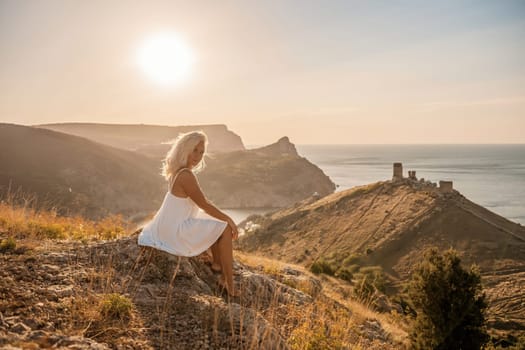 A woman is sitting on a hillside overlooking the ocean. She is wearing a white dress and has blonde hair. The scene is serene and peaceful, with the ocean in the background