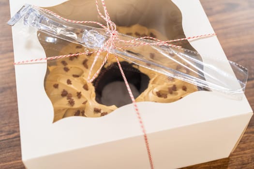 Carefully placing the gingerbread bundt cake, adorned with salted caramel frosting and gingerbread sprinkles, into a white paper bundt cake box with a clear window for gifting.