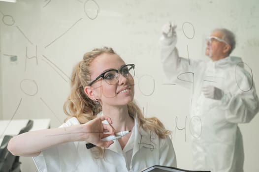 A woman chemist writes a formula on glass. An elderly Caucasian man in a protective suit is doing tests