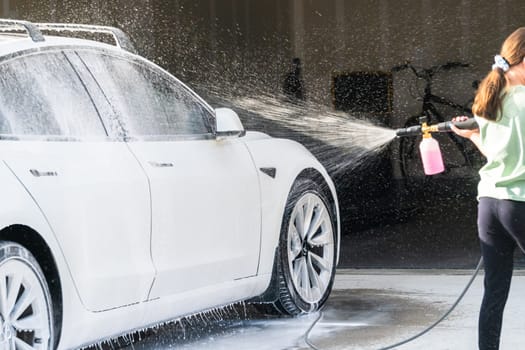 Denver, Colorado, USA-September 1, 2023-A young girl enthusiastically assists in washing the family's electric car in their suburban driveway.