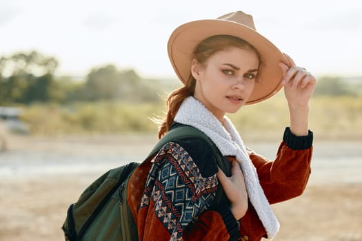 Woman in hat and backpack poses in field with car in background on sunny day