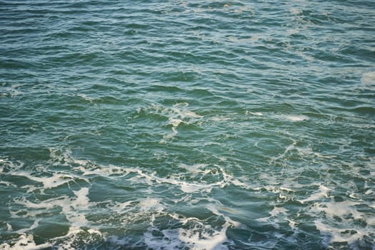 A closeup shot of the ocean with wind waves crashing onto the shore, creating a beautiful pattern of furrows in the water. The landscape is enhanced by cumulus clouds in the sky