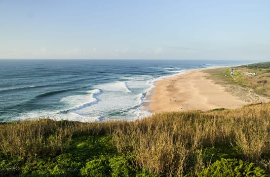 A breathtaking aerial view of a sandy beach and crystalclear ocean waters from a majestic cliff surrounded by lush greenery and coastal landforms. Nazare, Portugal