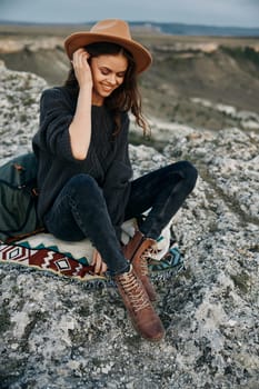 Woman in sweater and hat relaxing on rock with blanket on sunny day in nature