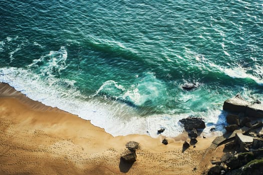 A breathtaking aerial view of a sandy beach and crystalclear ocean waters from a majestic cliff surrounded by lush greenery and coastal landforms. Nazare, Portugal