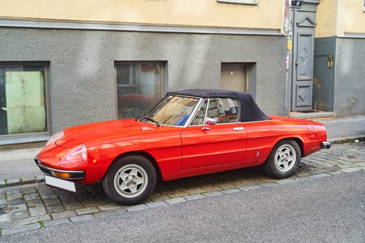 A red car with a black top is parked on the side of the road, with its automotive side marker lights and shiny wheels catching the sunlight