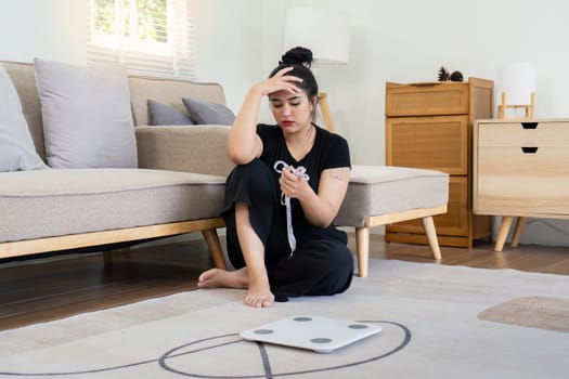 Stressed woman sitting on the floor holding a tape measure with a scale nearby, highlighting weight concern and anxiety