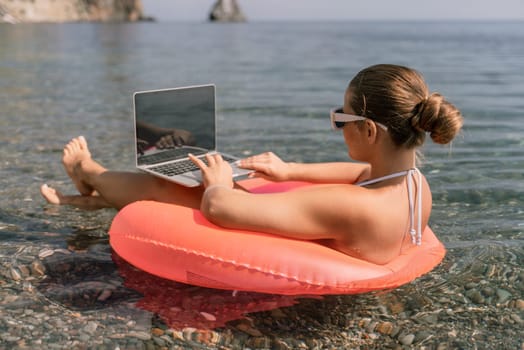 A woman is sitting on a pink inflatable raft in the water, using a laptop. Concept of relaxation and leisure, as the woman is enjoying her time by the water while working on her laptop