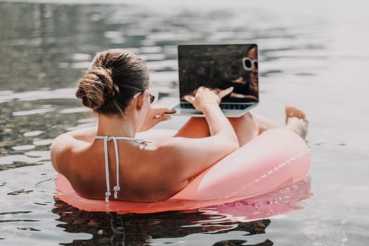 A woman is floating on a pink inflatable raft in the ocean while using a laptop. Concept of relaxation and leisure, as the woman enjoys her time in the water while working on her laptop