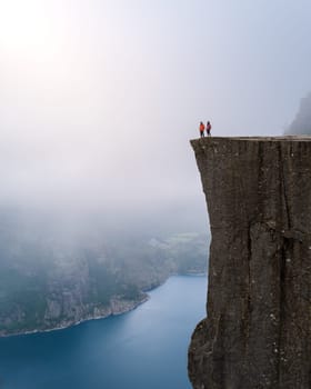 A breathtaking view from the Preikestolen cliff in Norway, with two figures standing at the edge looking out over the vast landscape. a couple of men and women hiking the Preikestolen, Norway