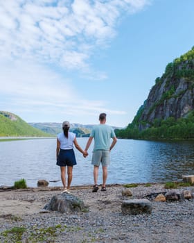 A couple stands hand-in-hand on the shore of a serene lake in Norway, gazing out at the surrounding mountains and clear blue sky.