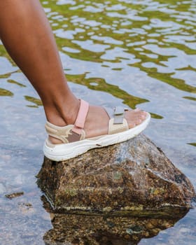 A foot adorned with a comfortable sandal rests on a smooth, gray stone partially submerged in clear, rippling water.