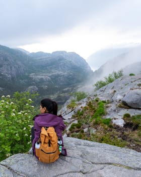 A woman sits on a rocky cliff overlooking a misty Norwegian fjord, her backpack beside her. The view is breathtaking, with the clouds rolling in and the mountains rising in the distance.