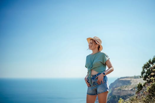 A woman wearing a hat and shorts stands on a cliff overlooking the ocean. She is enjoying the view and the fresh air